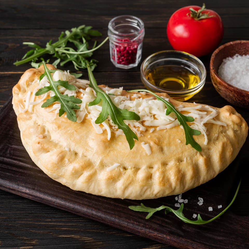 Brooklyn Pizzeria calzone topped with shredded cheese and arugula, alongside tomatoes, oil, and spices.