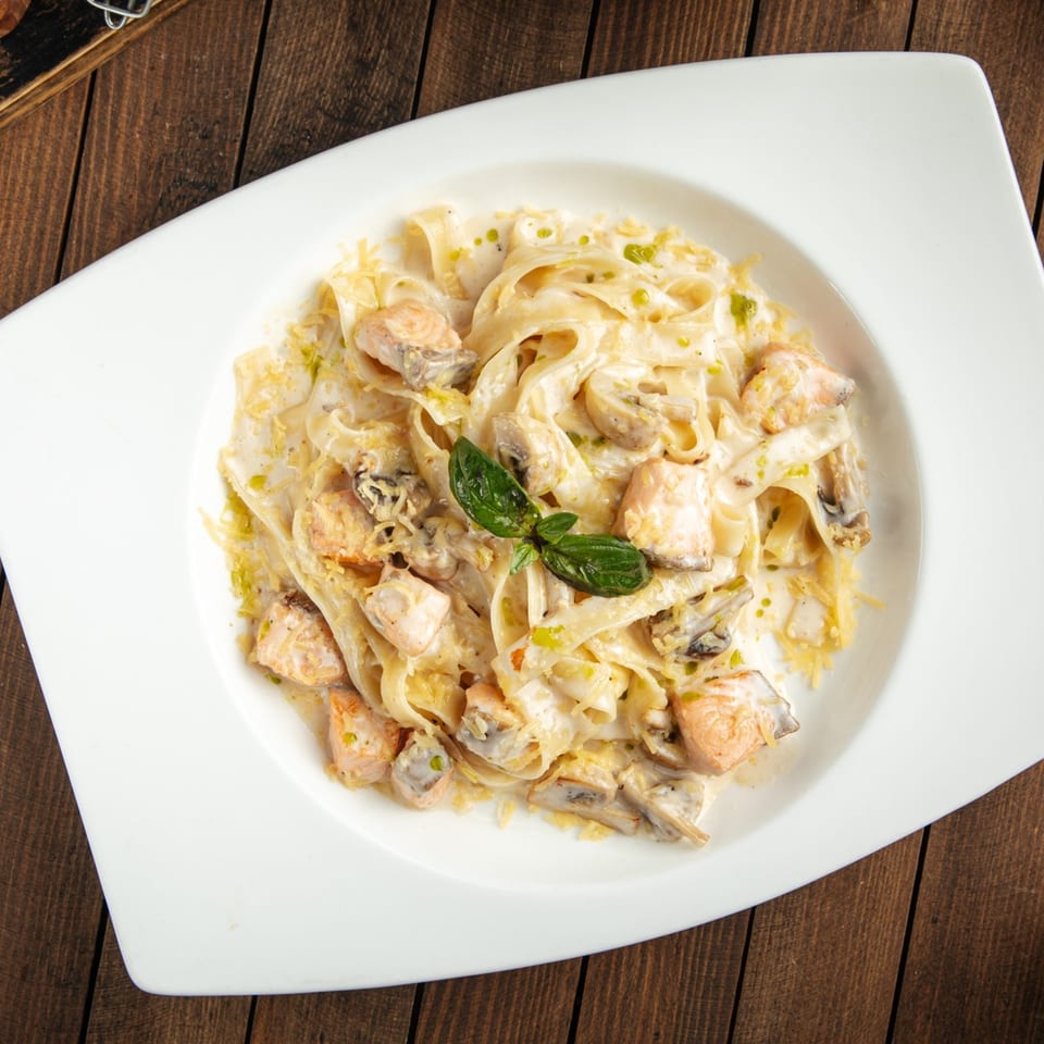 Creamy fettuccine pasta with salmon, mushrooms, and basil in a white bowl on a wooden table.