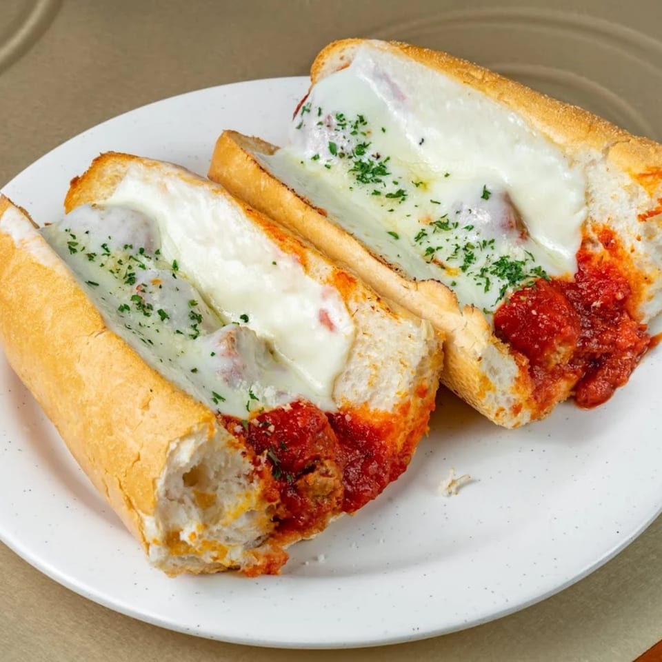 Two Brooklyn Pizzeria meatball subs with melted mozzarella, marinara sauce, and parsley on a white plate.